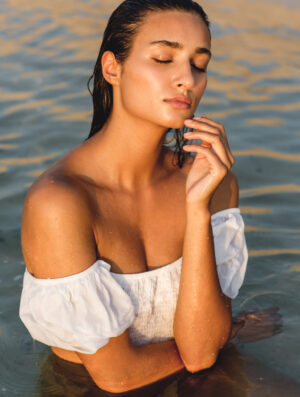 Portrait of exotic beautiful woman standing in the ocean water wearing a wet white sleeveless top during sunset.